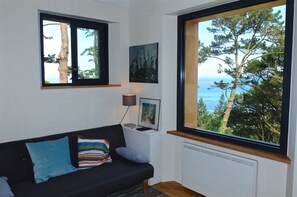 A modern living room with a black sofa, a small side table with a lamp, and framed pictures. There are two windows providing views of trees and the sea. - Kerpilu, listed *** house in a magical place facing the sea, along the GR34. (Plouguerneau)