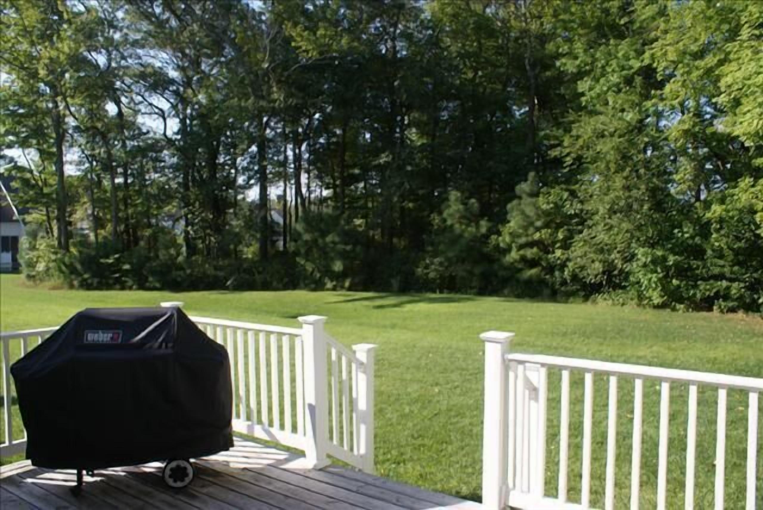 Een barbecue op een houten terras met witte relingen, omringd door een verzorgd gazon en bomen.