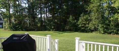 A barbecue grill on a wooden deck with white railings, surrounded by a well-maintained lawn and trees.