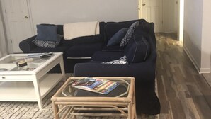 A living room with a dark blue sofa, a glass coffee table with magazines, a white side table, and a wooden floor. - Don't Miss Out on the 'Sunshine'; Unwind, Relax, Put Your Feet Up and enjoy! (Kill Devil Hills)