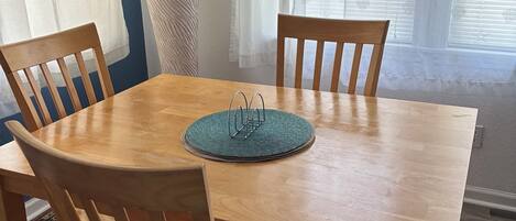 A dining area with a wooden table, four chairs, a woven placemat, a lamp, and a window with star-patterned curtains.