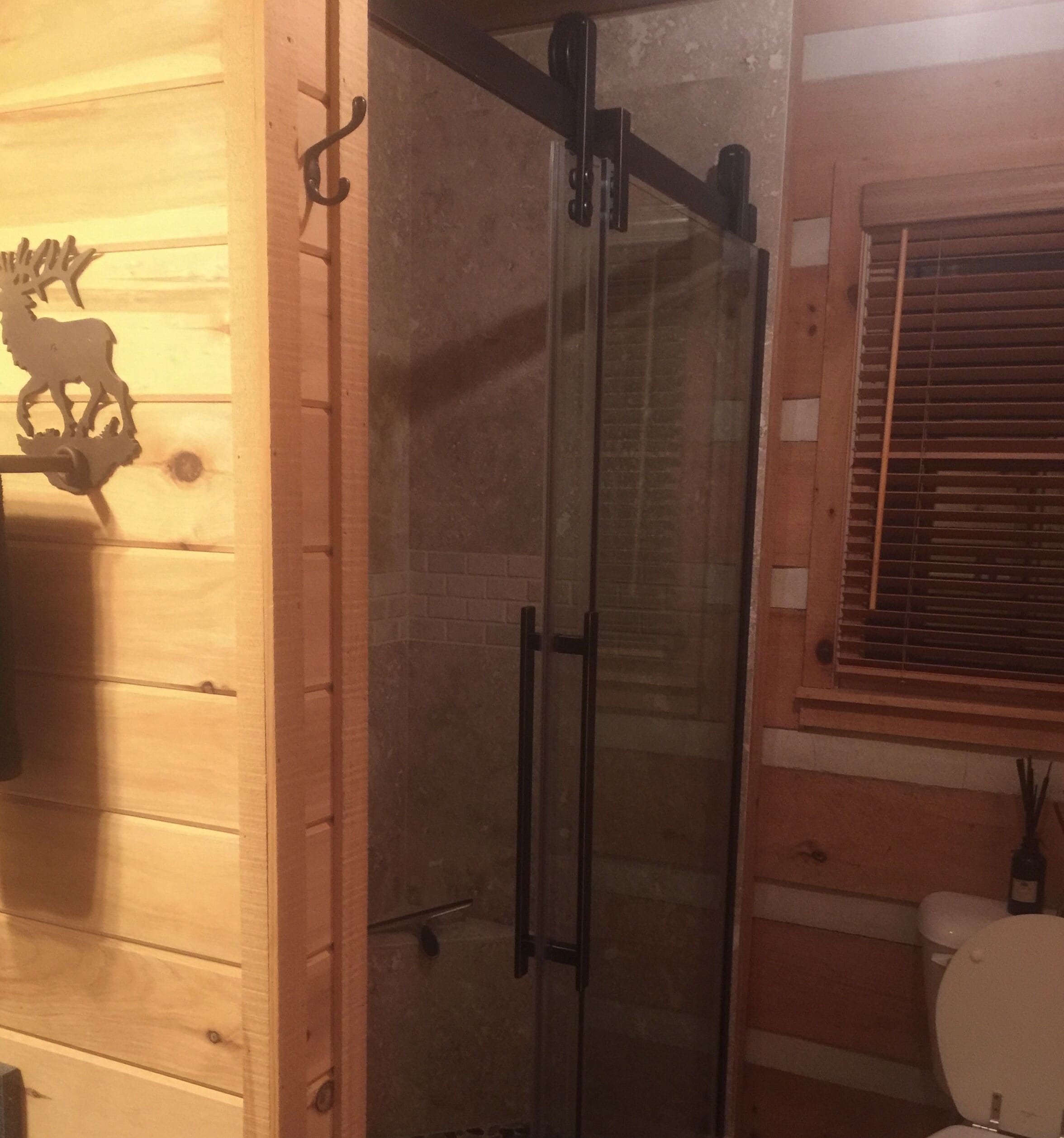 Ducha con puerta de vidrio, pared de madera con decoración de cabeza de ciervo y ventana con persianas.