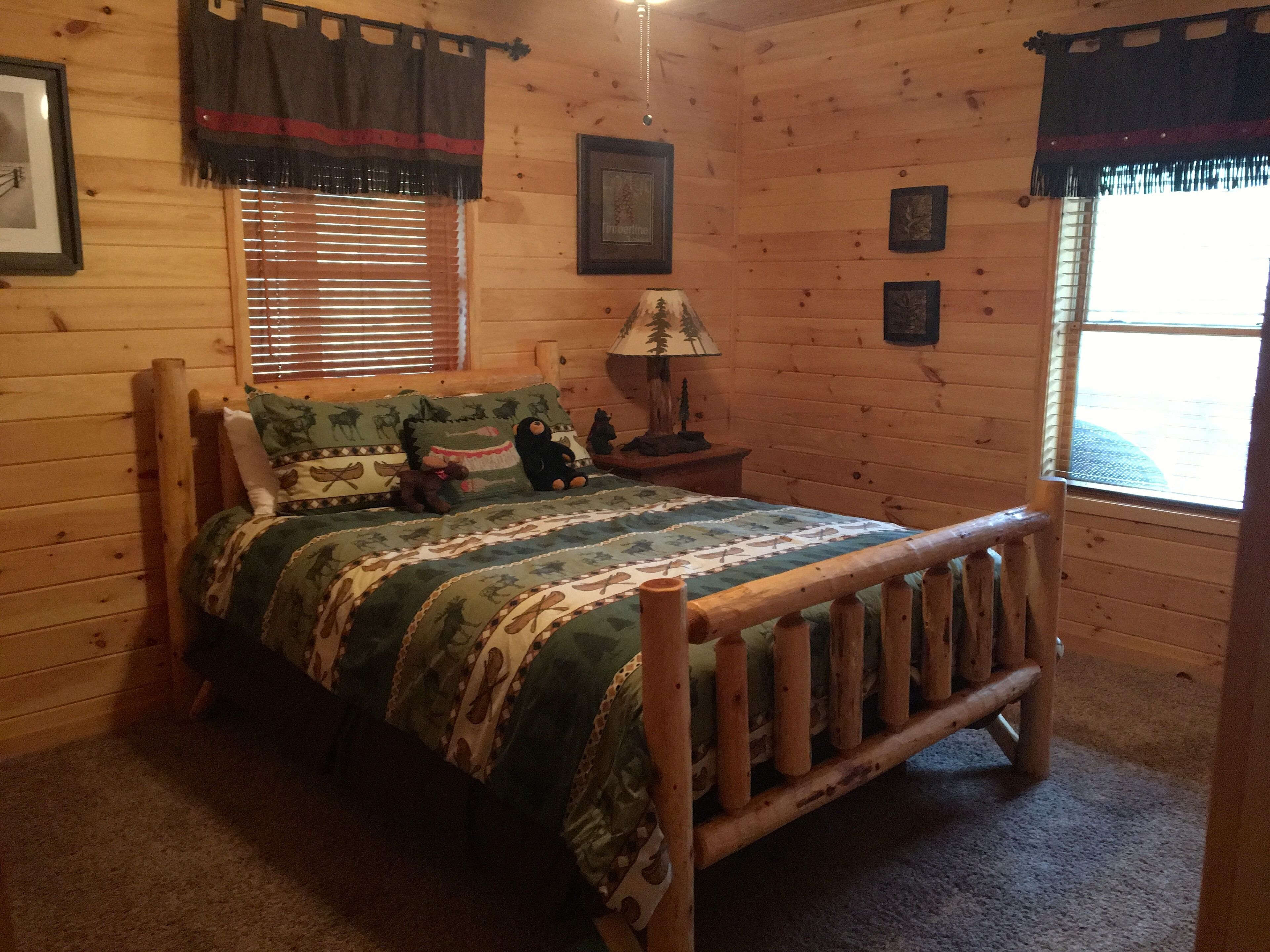 Um quarto de cabana rústica e aconchegante, com uma cama de madeira, uma mesinha de cabeceira com abajur e quadros decorativos nas paredes.