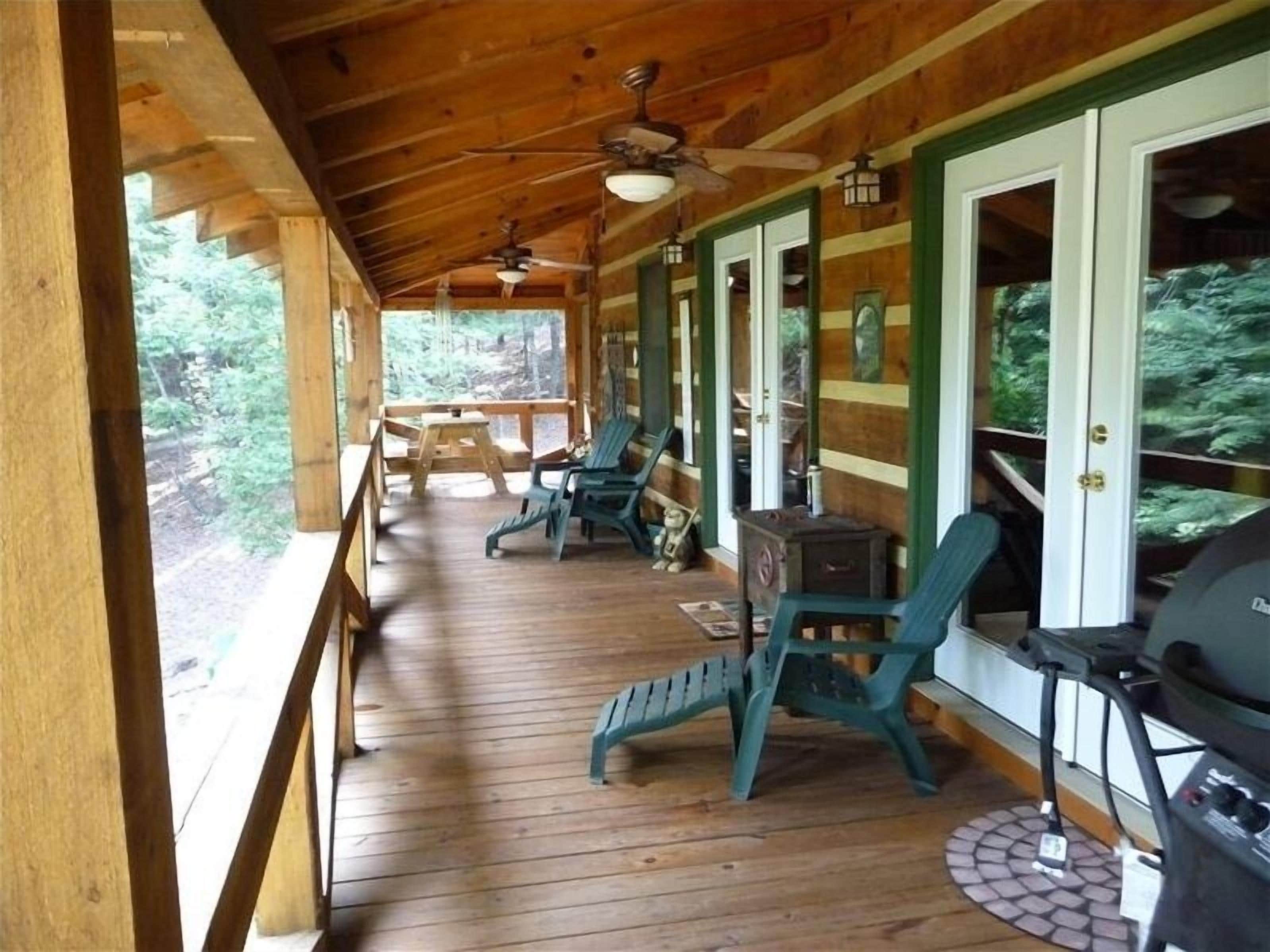 A covered porch with wooden flooring, green chairs, a grill, and a ceiling fan.