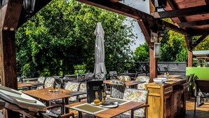 An outdoor patio with wooden tables and chairs, a bar area, and a view of lush greenery.