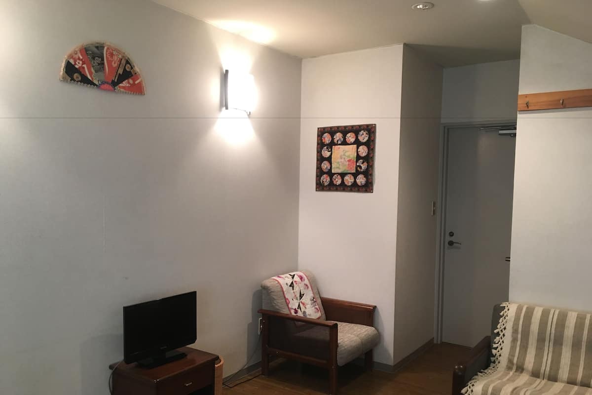 A room with a bed, a TV on a wooden stand, a chair, a wall-mounted light, and a door.