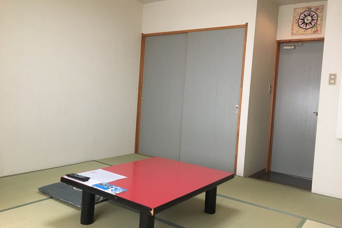A room with tatami flooring, a red table with papers, and two doors.