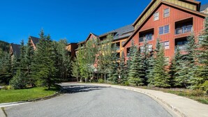 A multi-story building with a red exterior, surrounded by evergreen trees, and a curved driveway leading to the entrance. - 8884 | Springs, wood floors, steps to gondola! (Keystone)