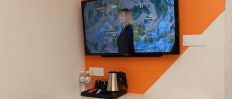 Habitación moderna con una silla blanca, un televisor de pantalla plana montado en una pared naranja, un pequeño escritorio blanco con una tetera y un cubo de basura.