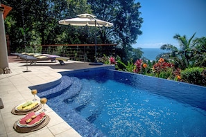 A swimming pool with a view of the ocean, surrounded by lush greenery and a lounge area with sunbeds and a table with drinks. - Perfect Location, Walk to Beach, Infinity Pool, Luxury Decor, Fast Free Wifi! (Dominical)
