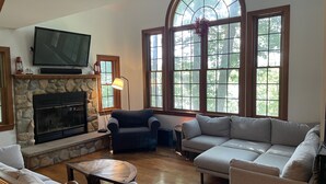 Una sala de estar con chimenea de piedra, televisor y ventanas amplias con vistas a árboles.