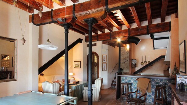 A spacious living area with a high ceiling, exposed wooden beams, a dining table, and a kitchen area with a tiled backsplash.