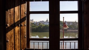 A view through a window with a wooden frame and a metal railing, overlooking a body of water and buildings in the distance.