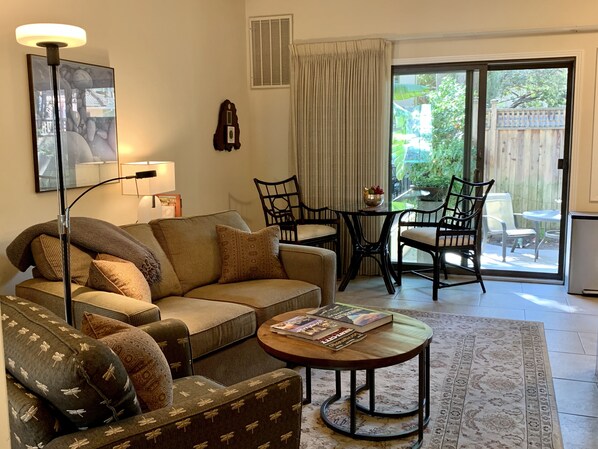 A cozy living room with a sofa, coffee table, and chairs, a painting on the wall, and a view of the backyard through a sliding glass door. - Quiet, Beautiful, East Sacramento Oasis - Walk to Coffee, Restaurants and Shops (SACRAMENTO)
