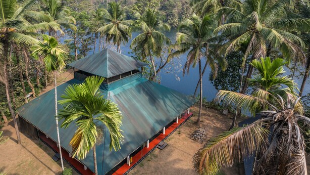 A bungalow with a green roof, surrounded by palm trees and a body of water. - Amritara Riverside Camp, Thattekad (Kothamangalam)