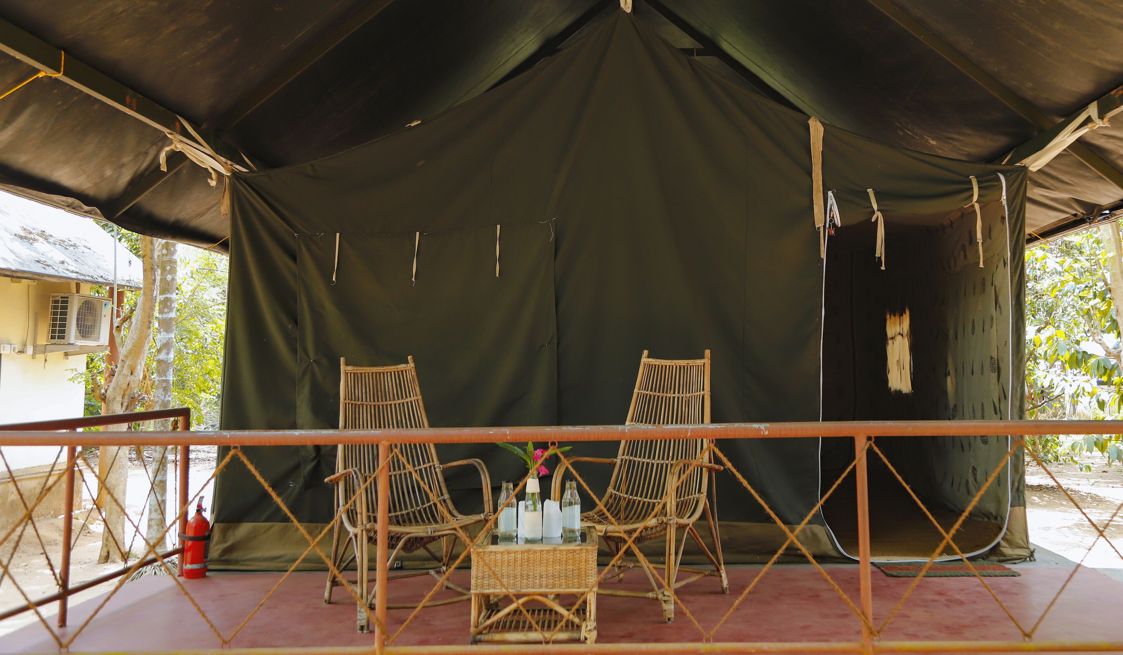 A covered outdoor seating area with two wicker chairs and a small table, a fire extinguisher, and a tent in the background.