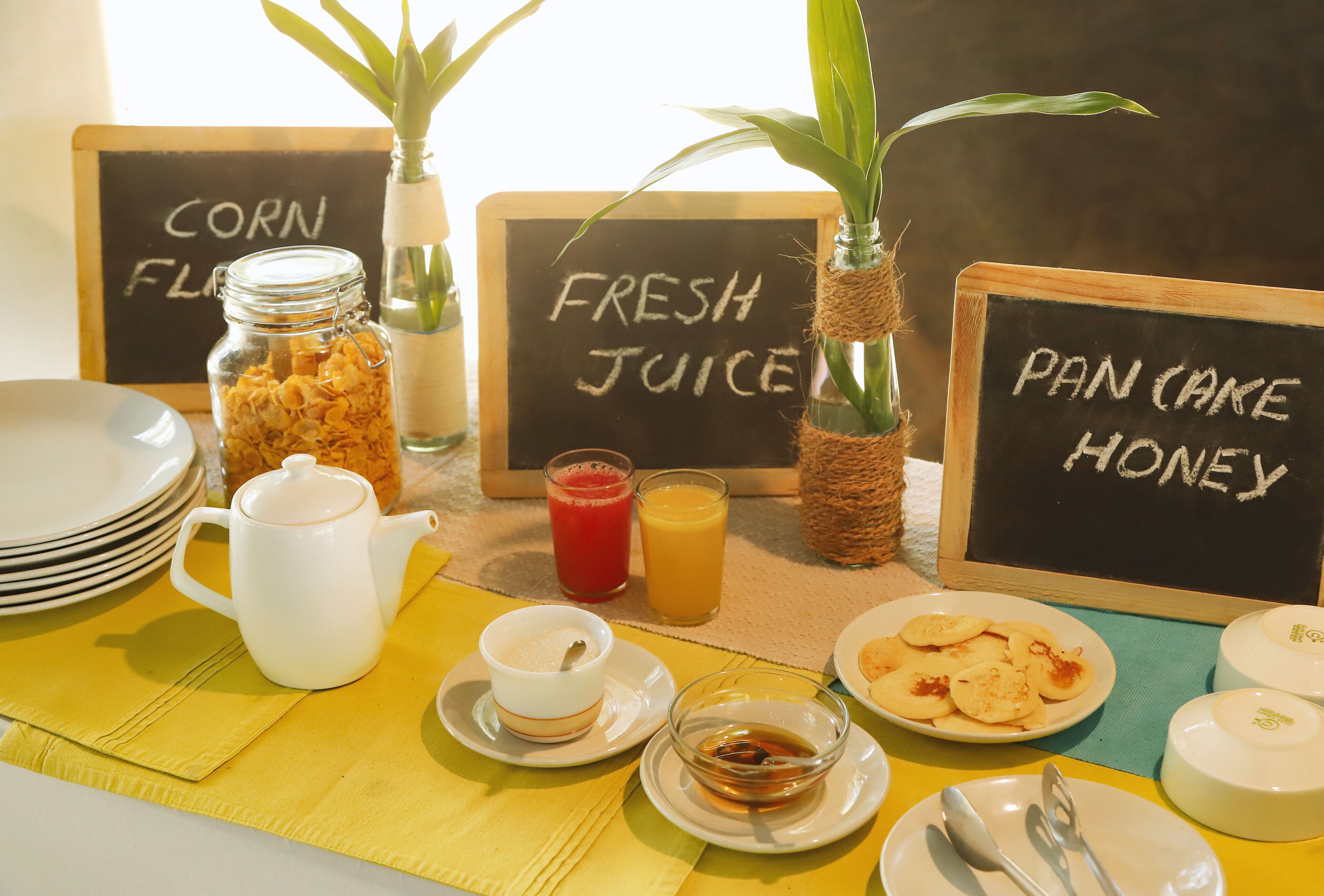 A breakfast setup with a white teapot, cups, and a plate of pancakes. There are also signs indicating 'Corn Flakes', 'Fresh Juice', and 'Pan Cake Honey'.