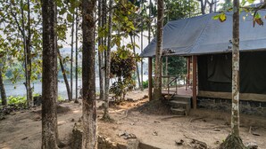 A rustic cabin with a covered porch overlooking a lake, surrounded by trees. - Amritara Riverside Camp, Thattekad (Kothamangalam)