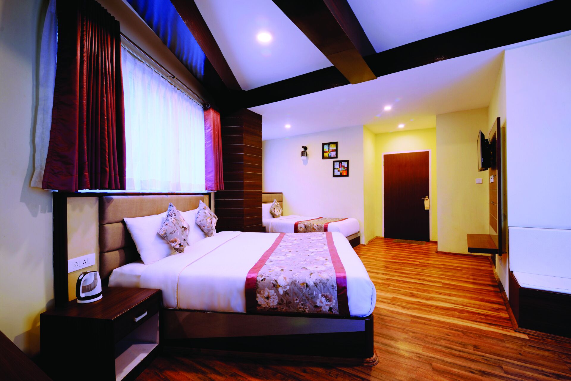 A hotel room with a large bed, a bedside table with a kettle, a wooden floor, and a door leading to another room.