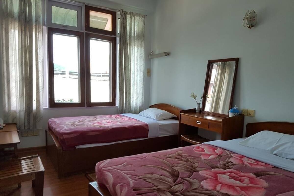 Quarto com duas camas, uma escrivaninha de madeira, um espelho e uma janela com cortinas.