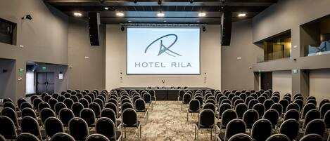 Un salón de conferencias con filas de sillas orientadas hacia una pantalla grande que muestra el logotipo del Hotel Rila.
