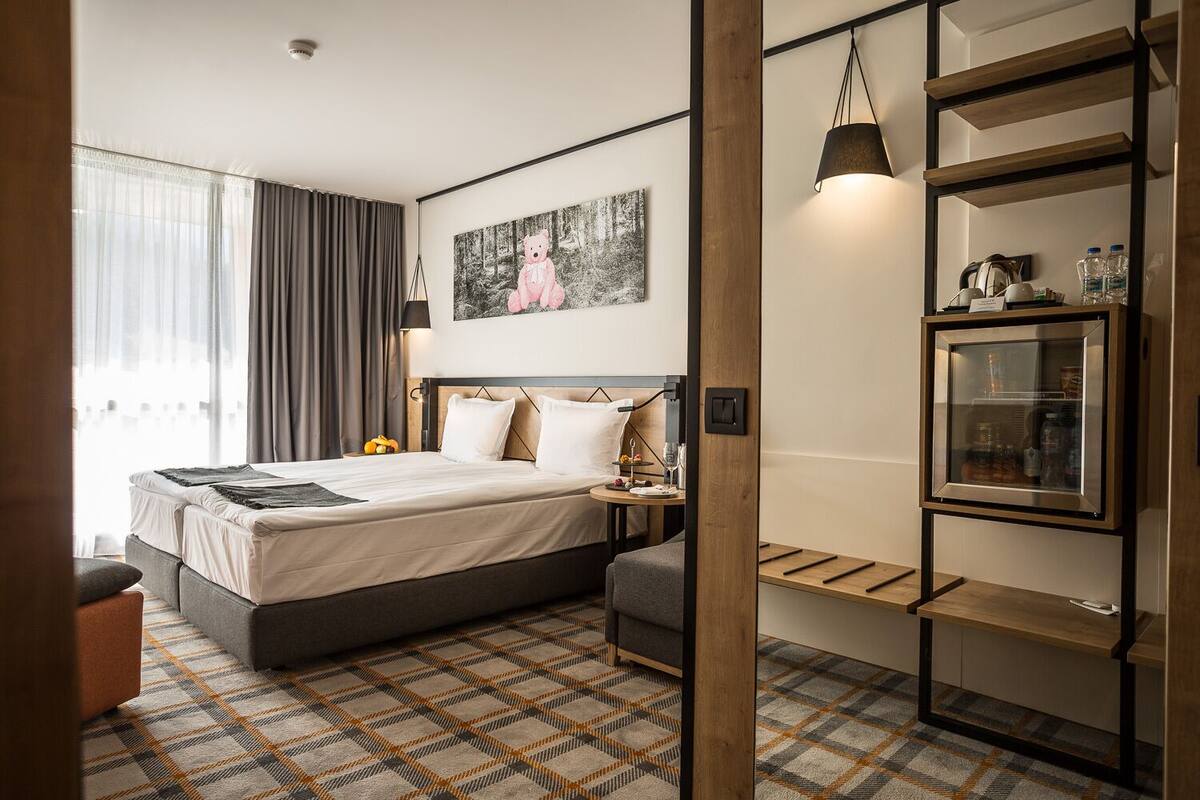 A modern hotel room with a large bed, a bedside table with a lamp, a small seating area, and a built-in shelf with a refrigerator.