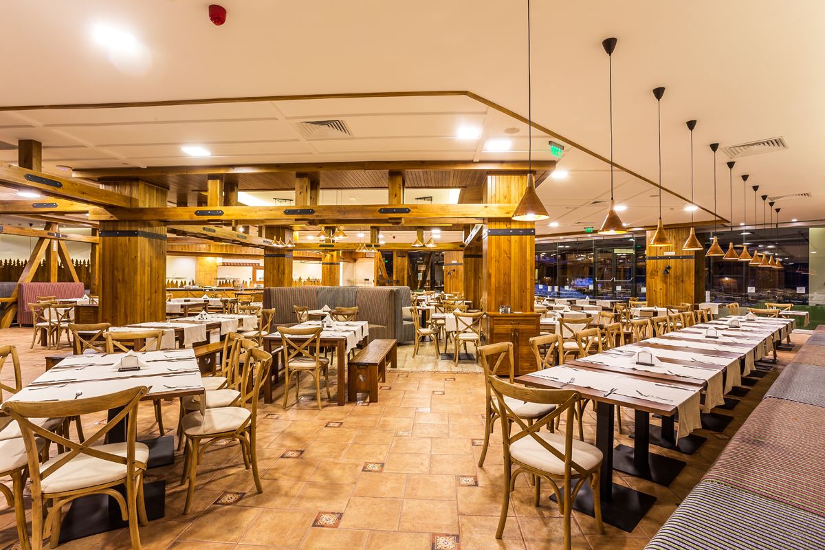 Een ruim restaurant met lange gedeelde tafels en houten stoelen, met een hoge plafond met zichtbare balken en hanglampen.