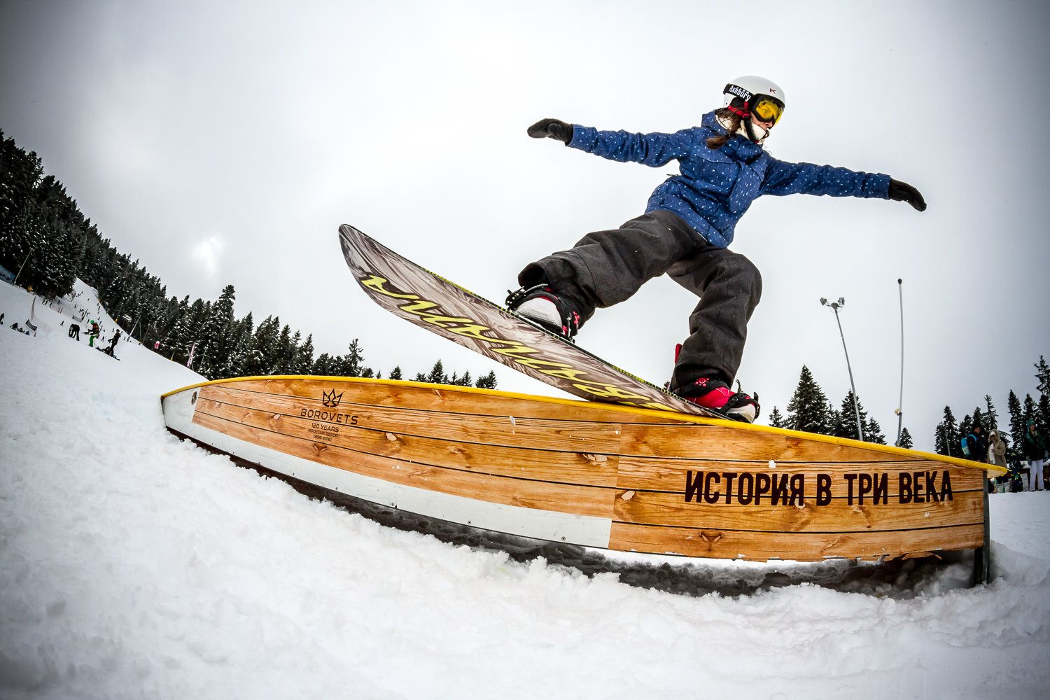 A snowboarder performing a trick on a wooden rail with text in Cyrillic script.