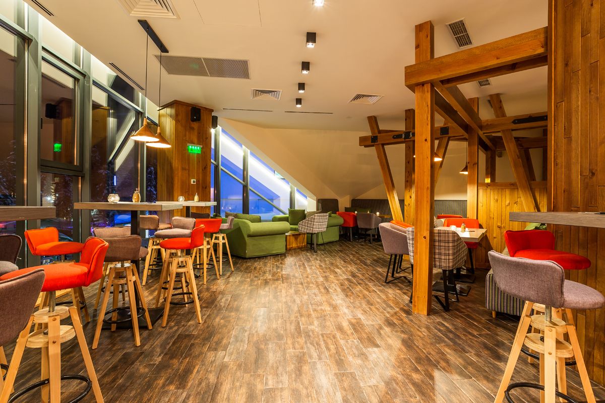 A modern interior with wooden beams, a mix of seating options including bar stools and sofas, and large windows providing natural light.