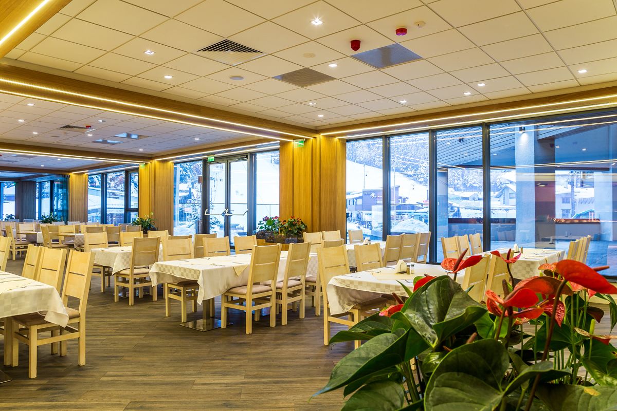 A spacious dining area with multiple tables and chairs, large windows offering a view of a snowy landscape, and a warm ceiling design.