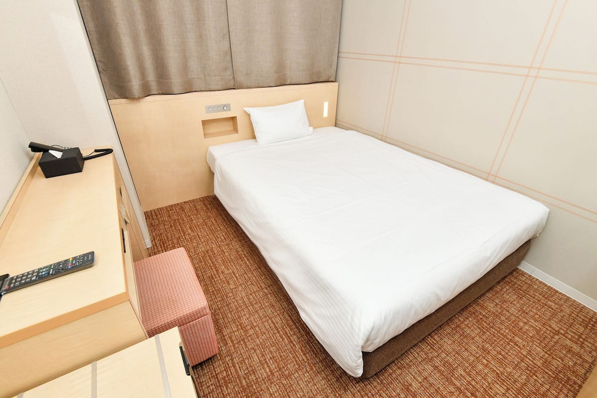 Una habitación de hotel con una cama individual, un escritorio con teléfono y control remoto, y una alfombra marrón.