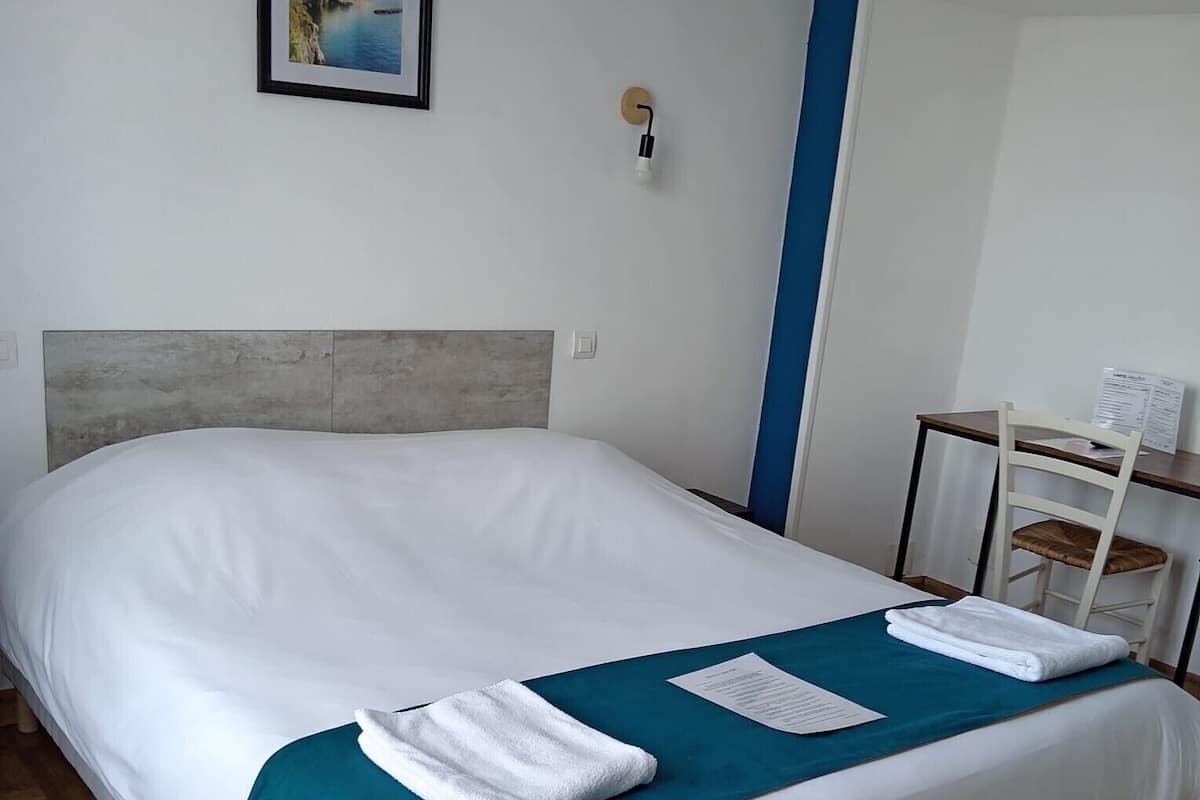 Una cama bien tendida con sábanas blancas y una manta color turquesa, un cuadro en la pared y una pequeña mesa con una silla a la derecha.