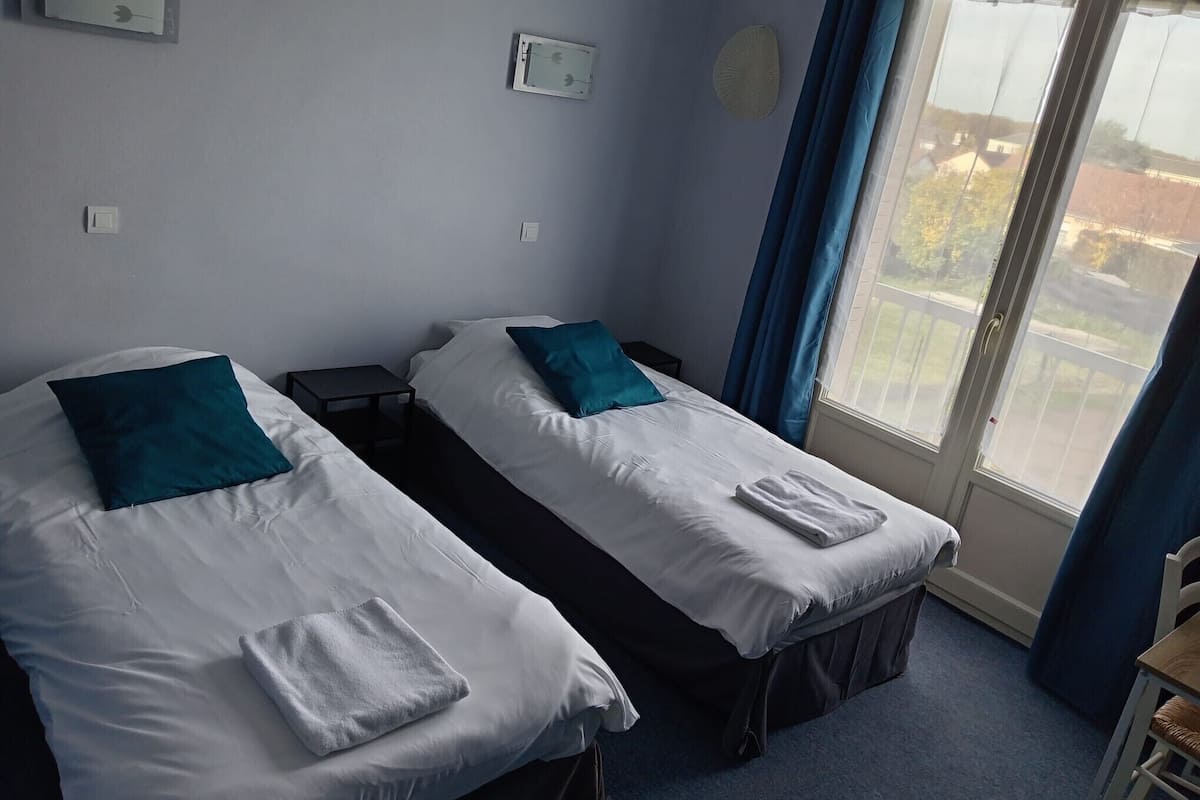 Habitación de hotel con dos camas individuales, una mesita, una silla y una ventana con cortinas.