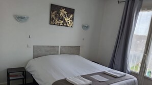 A neatly made bed with white linens and a brown throw blanket, a small bedside table with a book, and a painting of tropical flowers on the wall.