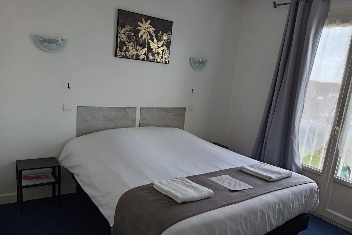 Una cama bien hecha con sábanas blancas y una manta marrón, una mesita de noche con un libro, y un cuadro con flores tropicales en la pared.