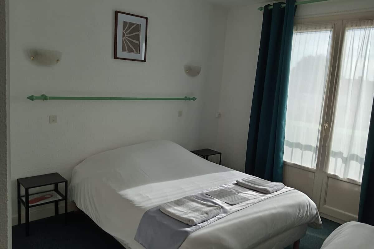 Habitación de hotel con una cama individual, una mesita de noche pequeña, un cuadro enmarcado en la pared y una ventana con cortinas.