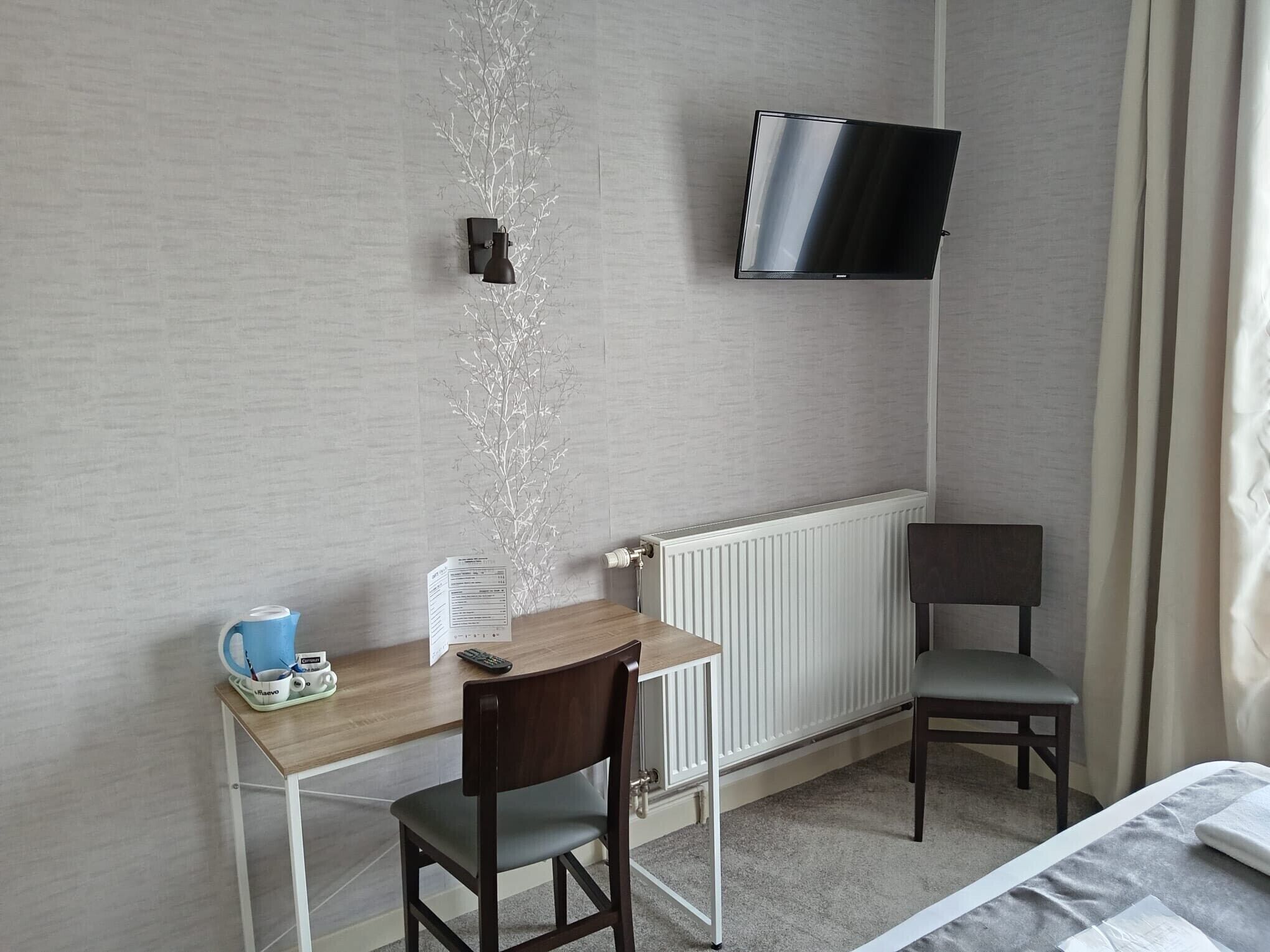 Une chambre d’hôtel avec un bureau en bois, une chaise, un téléviseur fixé au mur, un radiateur et un lit recouvert d’une couverture grise.