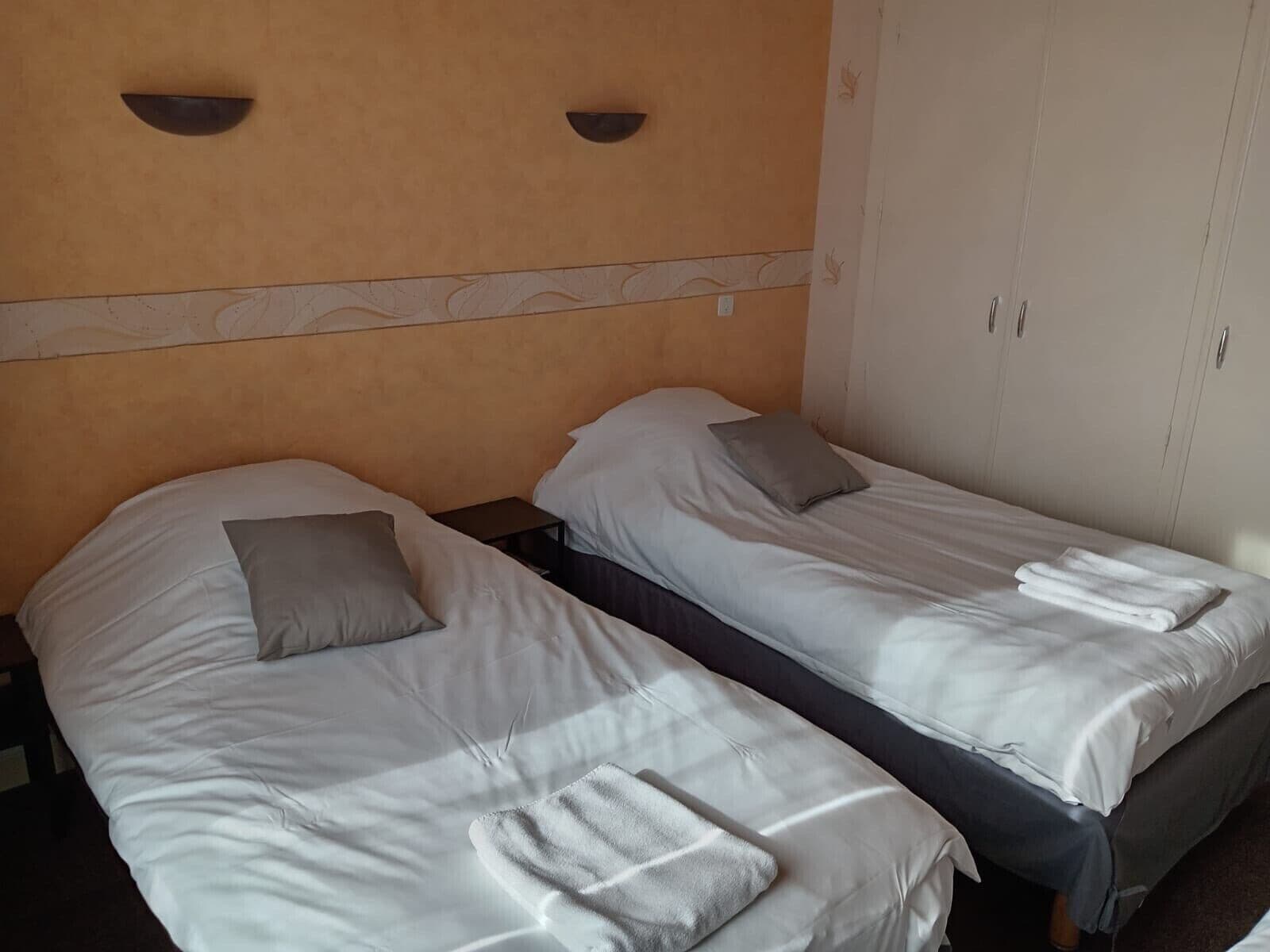 A hotel room with two single beds, each with a pillow and a folded towel. There is a nightstand between the beds, and a wardrobe with two doors to the right.
