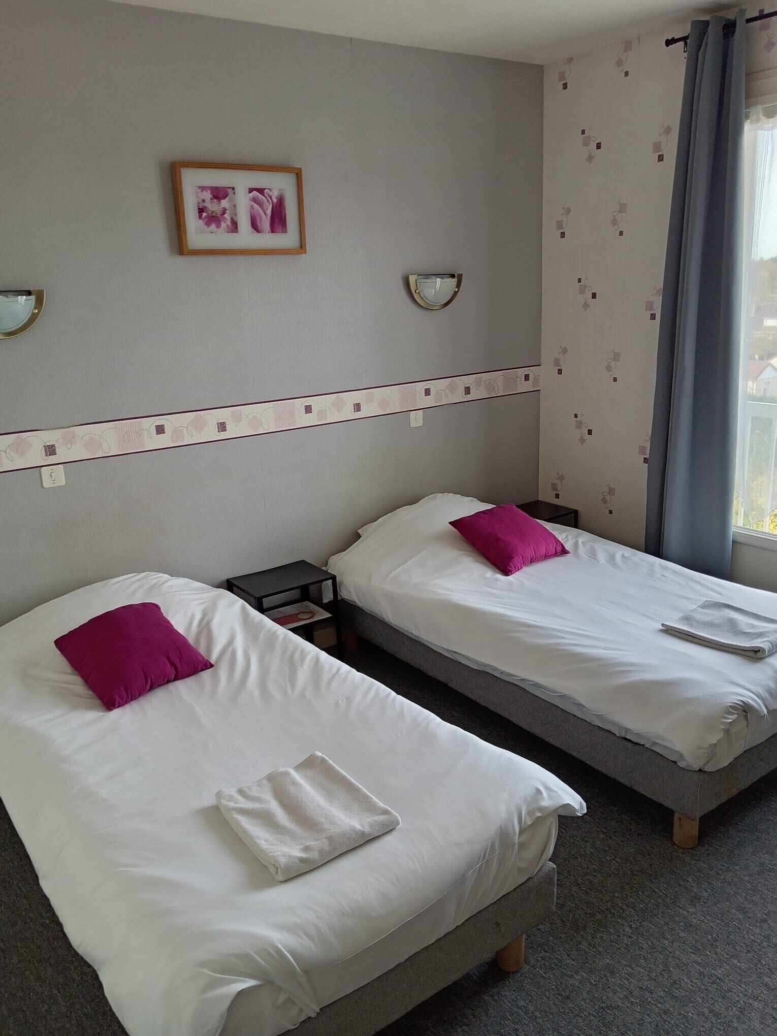 Una camera d'albergo con due letti singoli, un comodino con una lampada, un quadro appeso al muro e una finestra con tende.