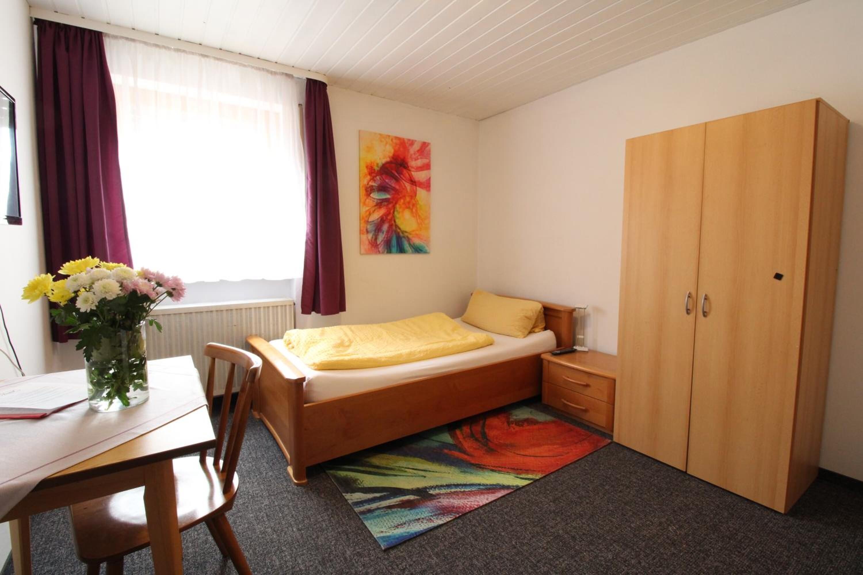 Una camera da letto con un letto, un armadio in legno, una scrivania con una sedia, un vaso con fiori e un quadro appeso al muro.