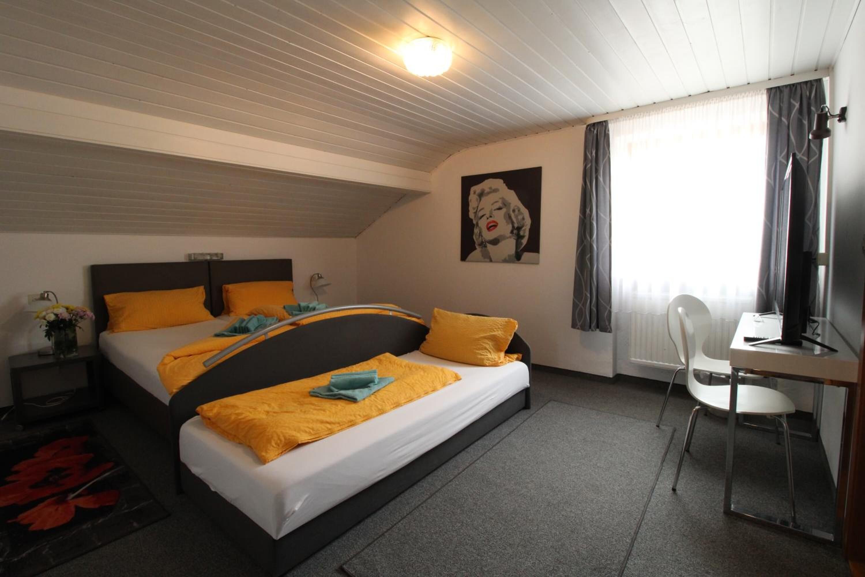 Ett sovrum med två sängar, ett skrivbord, en stol, ett fönster med gardiner, en tavla på väggen och en taklampa.