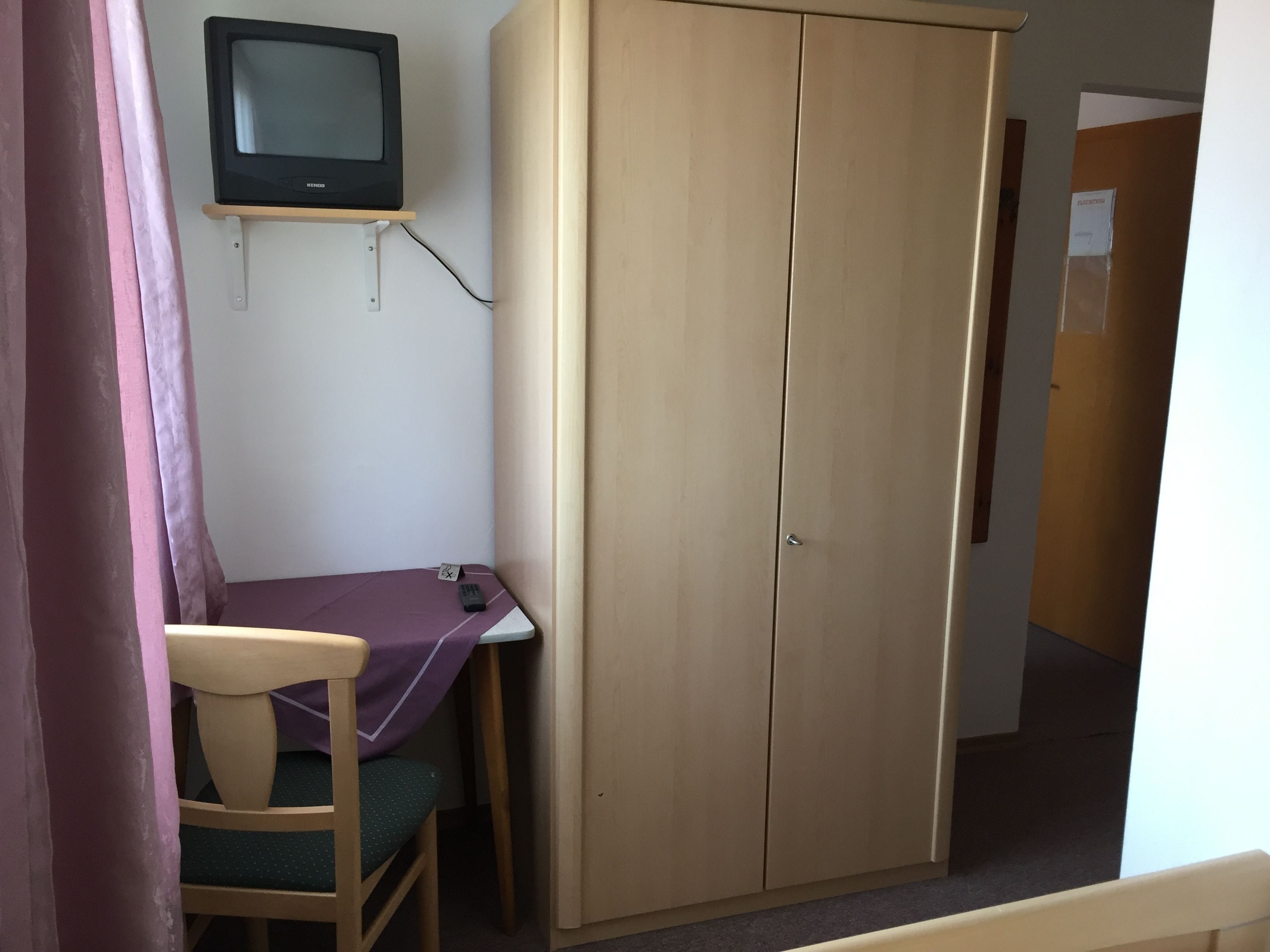 Ein Zimmer mit einem Holzschrank, einem kleinen Tisch mit lila Tischdecke, einem Stuhl und einem Fernseher auf einem Schrank.