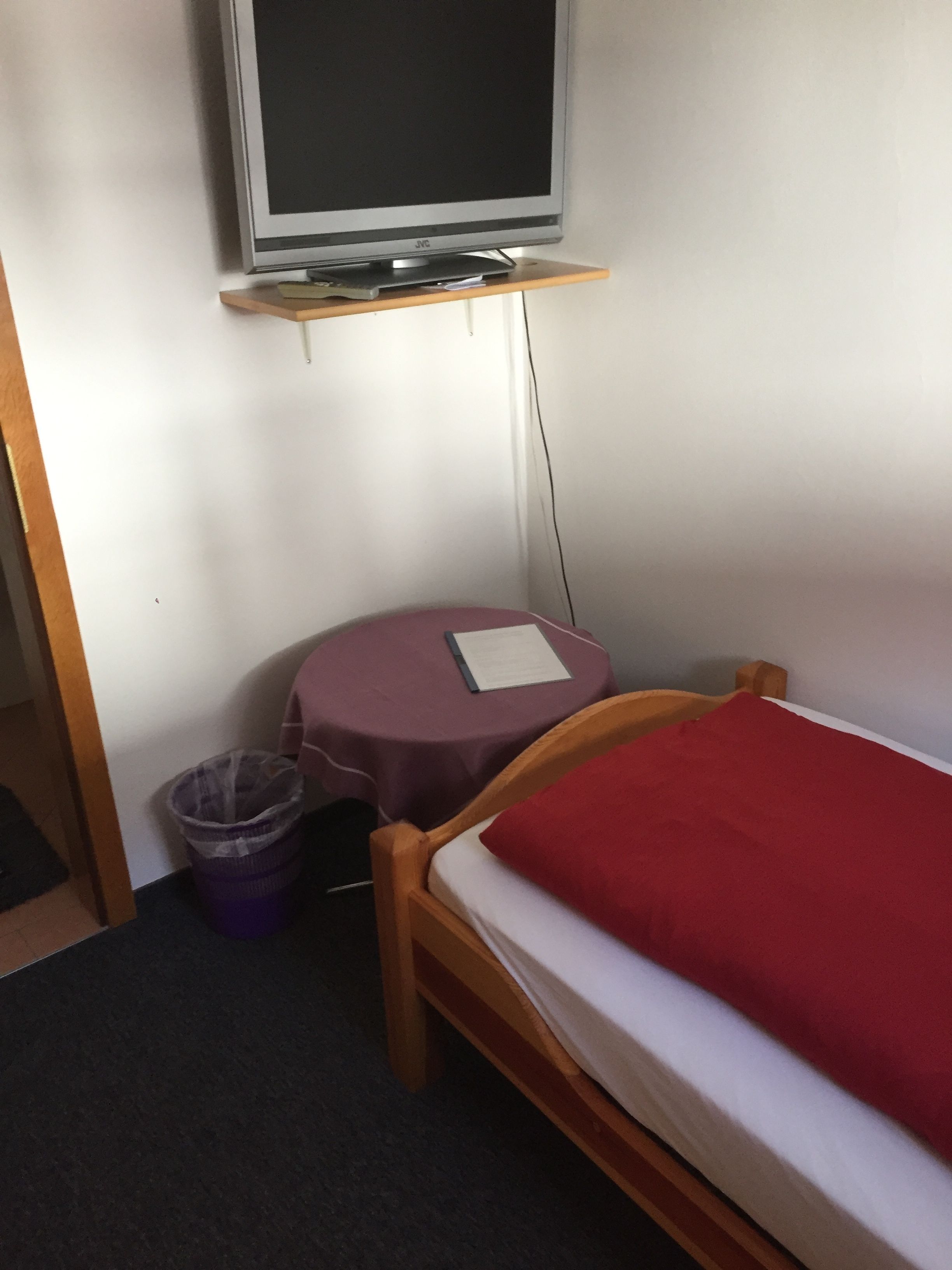 Una habitación pequeña con una cama individual, una mesa, una silla, un televisor sobre un soporte y un cubo de basura.