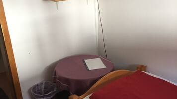 Una habitación pequeña con una cama individual, una mesa, una silla, un televisor sobre un soporte y un cubo de basura.