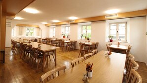 A spacious dining area with multiple wooden tables and chairs, large windows with curtains, and potted plants. - Zur Auszeit (Traunreut)