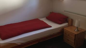 Une petite chambre avec un lit simple, un cadre en bois, un oreiller rouge, une table de chevet en bois avec une lampe et un radiateur.