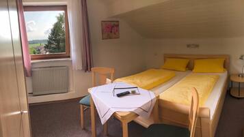 A bedroom with a bed, a table with a remote control, a chair, a window with curtains, and a radiator.