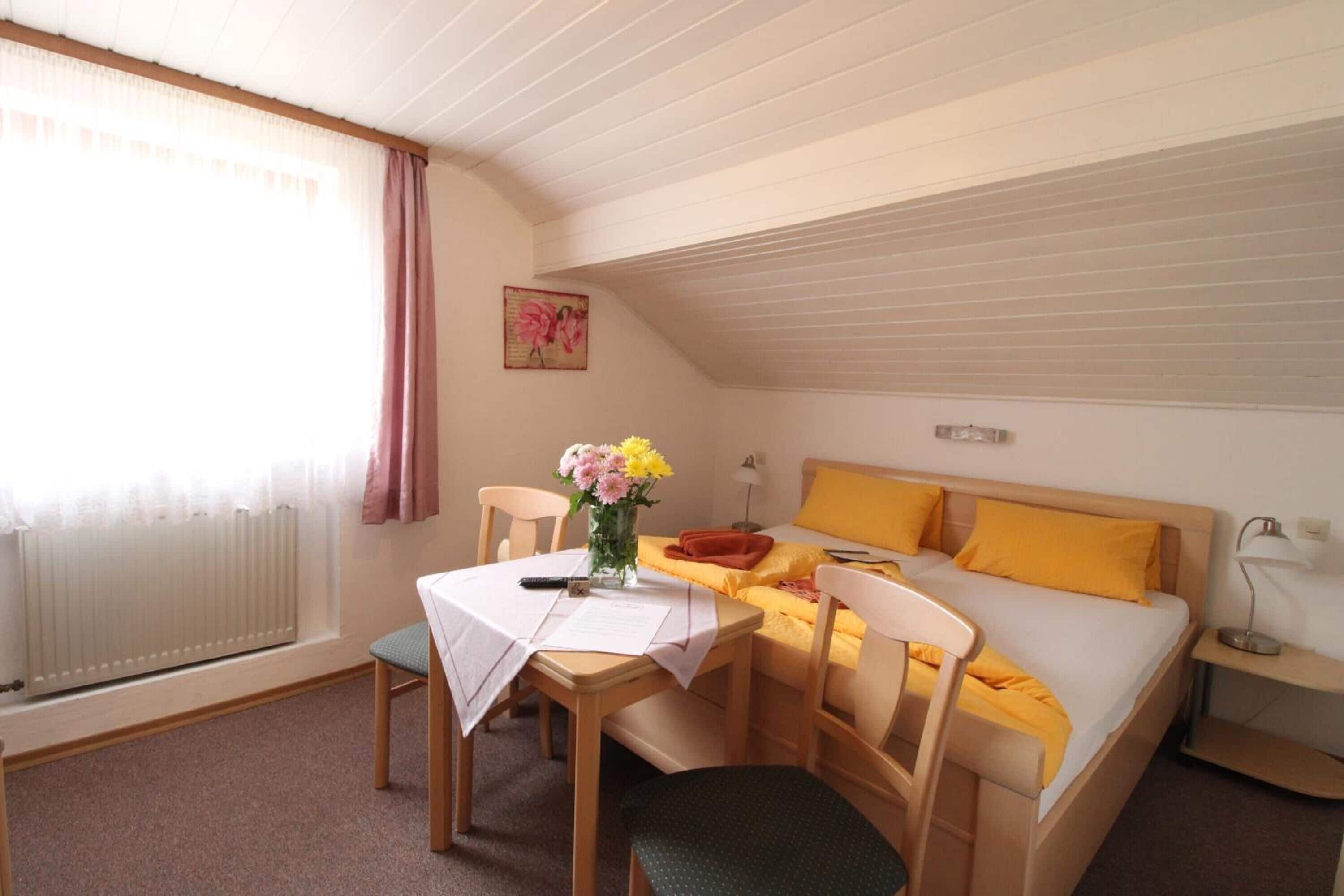 A cozy room with a bed, a table with chairs, a vase with flowers, and a window with curtains.