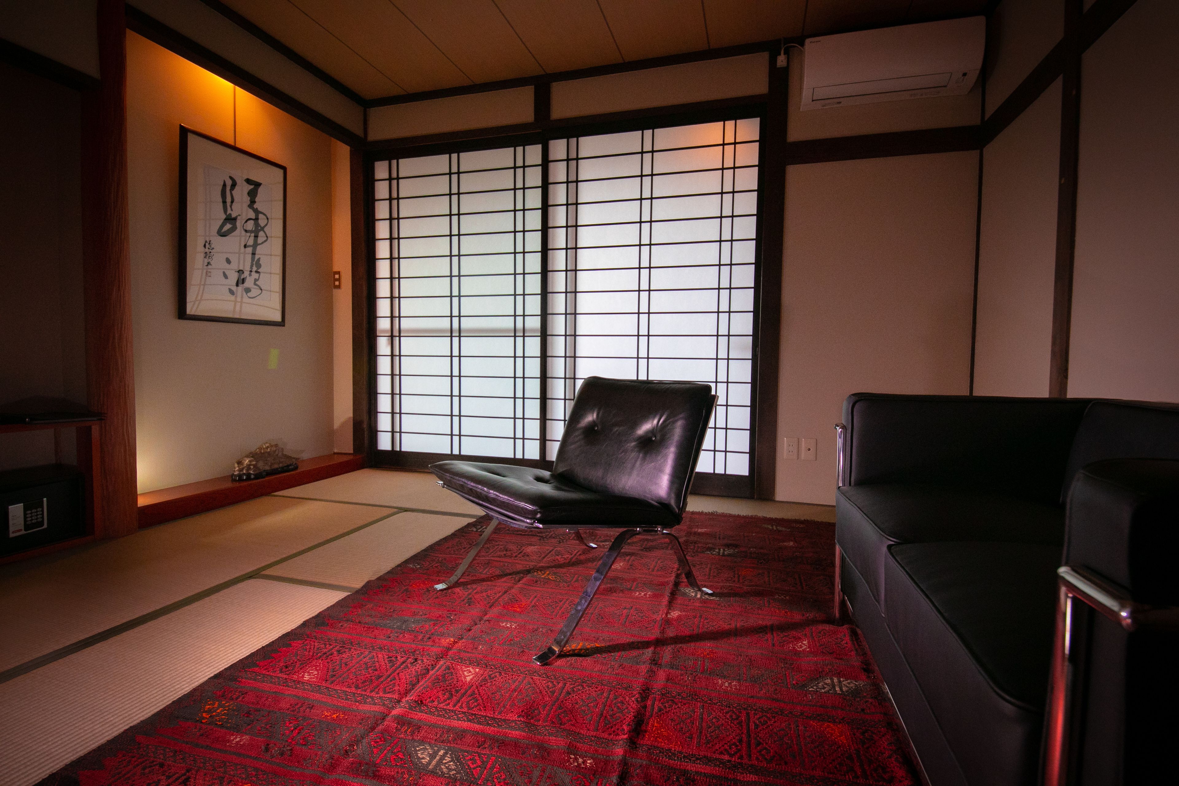 Una stanza tradizionale giapponese con pavimento in tatami, una poltrona in pelle nera, un divano nero e un paravento shoji con calligrafia.