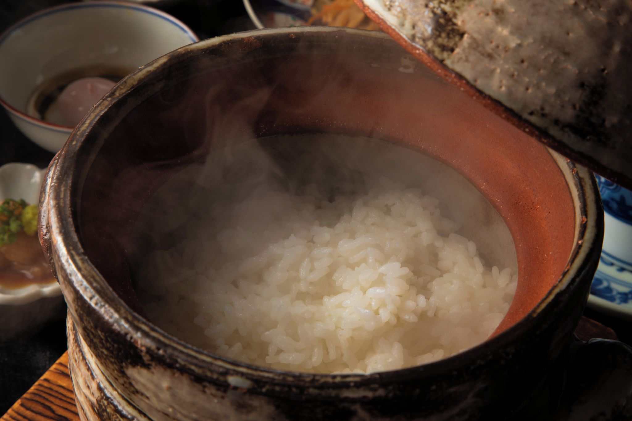 A traditional clay pot filled with steaming white rice, accompanied by a small bowl of sauce and a piece of bread.